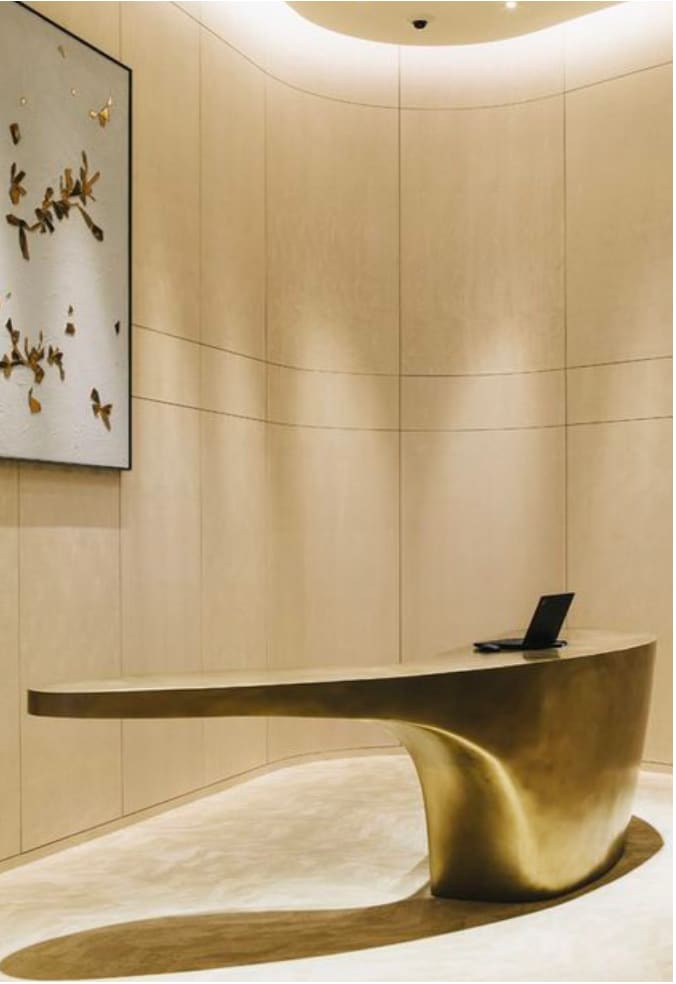 Curved host station inside a City Walk cafe, with warm beige stone-clad walls, a sculptural cantilevered brass counter, a recessed cove-lit ceiling and a polished terrazzo floor.